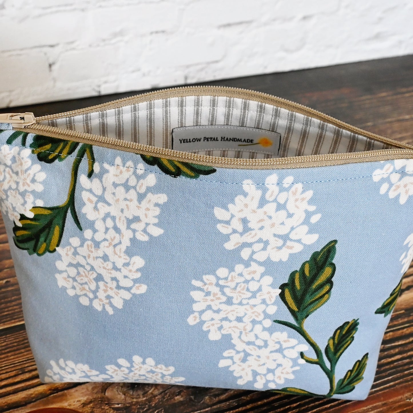 Small blue hydrangea accessory pouch made from Rifle Paper Co fabric. Zips at the top and is lined in a grey and cream striped cotton. Made in Canada by Yellow Petal Handmade.